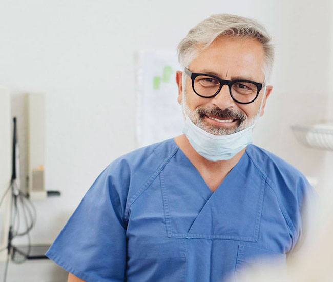 Zahnarzt in seiner Praxis mit dunkler Brille und grauen Haaren lächelt