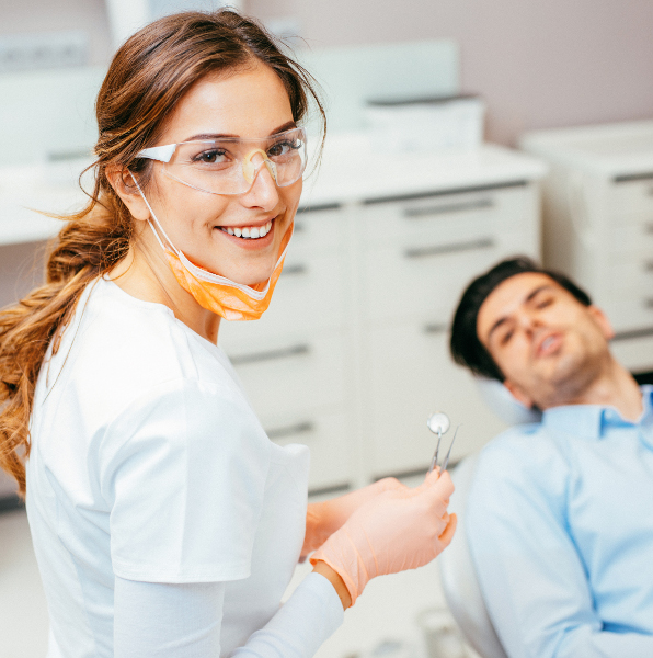 Zahnärztin lächelt in die Kamera, Patient liegt hinter ihr auf dem Behandlungsstuhl
